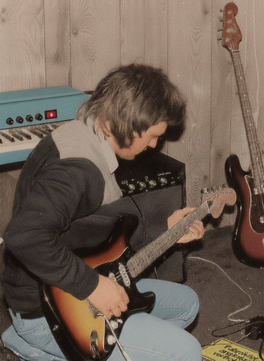 Che Guitare macht im Übungsraum einen Soundcheck mit seiner Gitarre, die er mit einem selbstgebauten Verstärker spielt. Im Hintergrund ist außerdem ein ebenfalls selbstgebautes Tasteninstrument zu sehen.